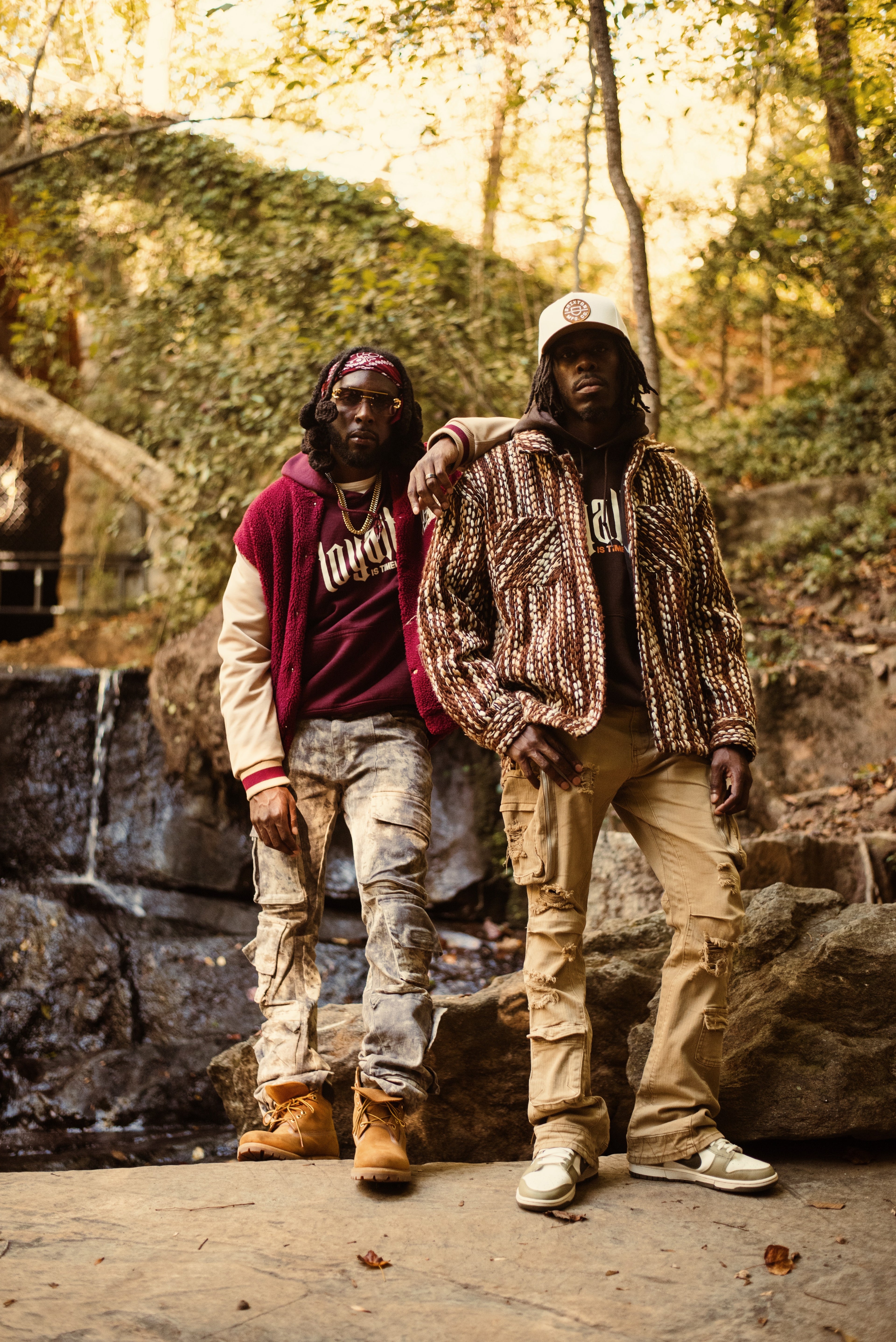 Two individuals standing outdoors near a waterfall with a natural background